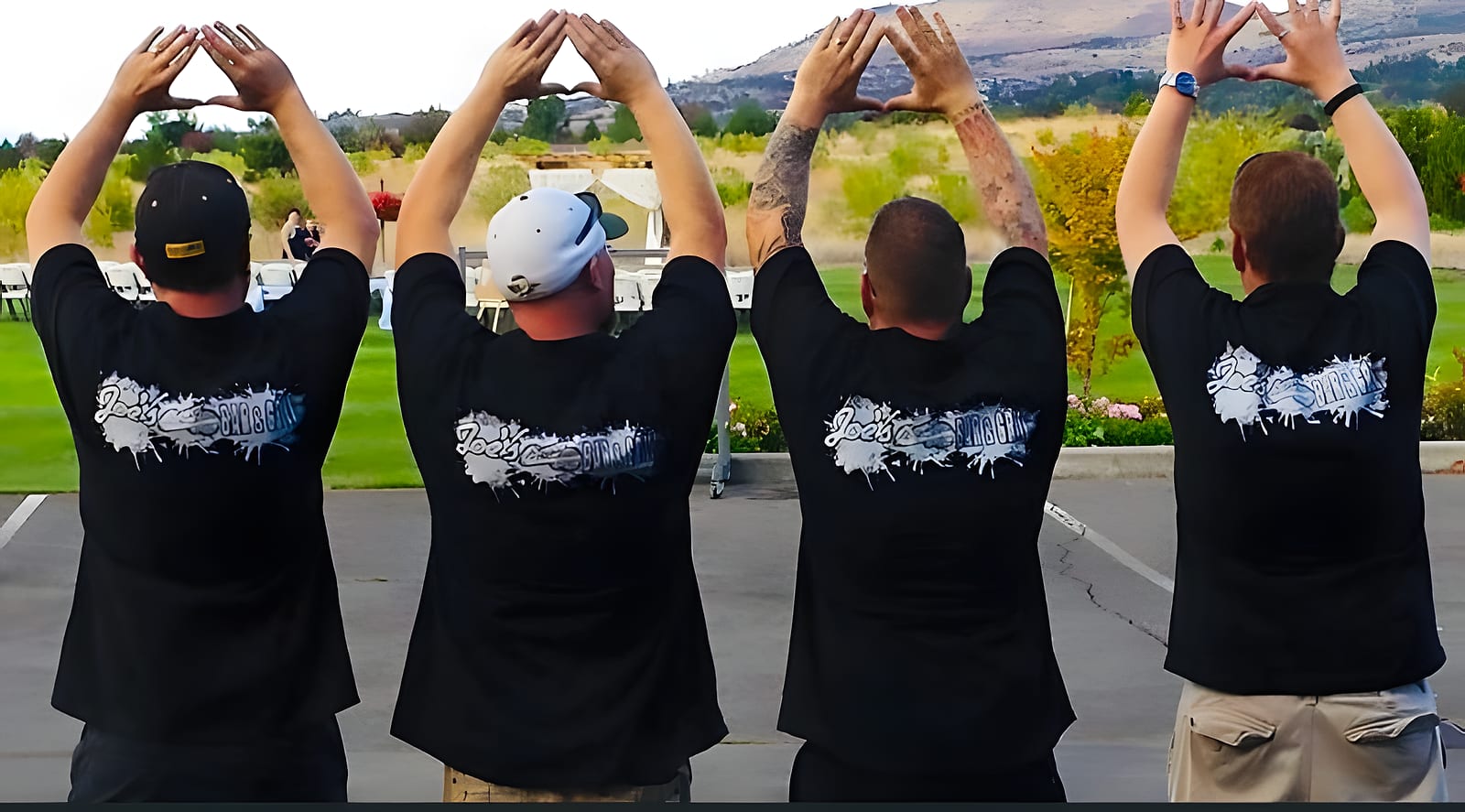 Four men in matching black bar T-shirts seen from behind, arms raised overhead